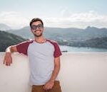 Young man in sunglasses leaning on a balcony with a scenic city view in the background.